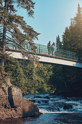Buy stock photo Shot of a young couple standing on a bridge surrounded by nature