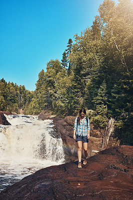 Buy stock photo Shot of an attractive young woman walking next to a rocky river and waterfall