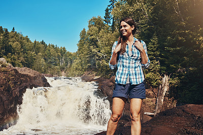 Buy stock photo Shot of an attractive young woman standing next to a rocky river and waterfall