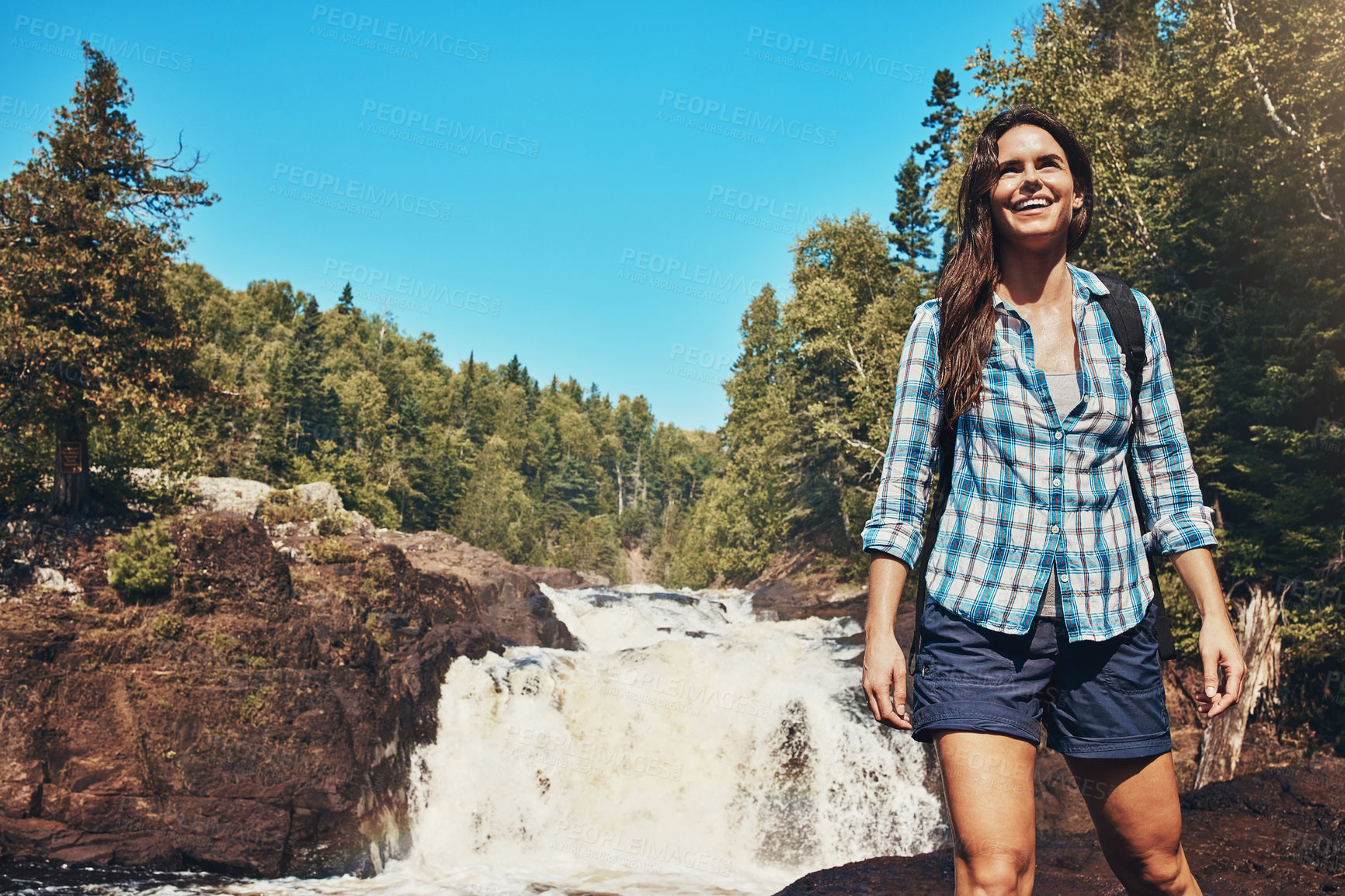 Buy stock photo Shot of an attractive young woman standing next to a rocky river and waterfall
