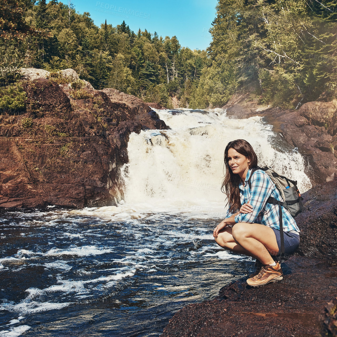 Buy stock photo Shot of an attractive young woman crouching next to a rocky river and waterfall