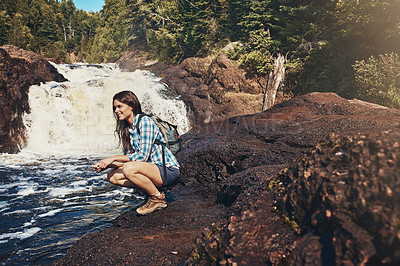 Buy stock photo Shot of an attractive young woman crouching next to a rocky river and waterfall