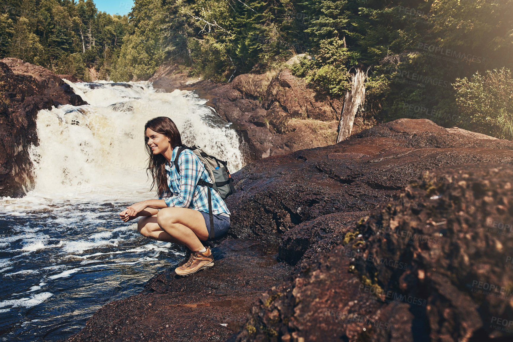 Buy stock photo Shot of an attractive young woman crouching next to a rocky river and waterfall