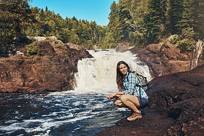 Buy stock photo Shot of an attractive young woman crouching next to a rocky river and waterfall