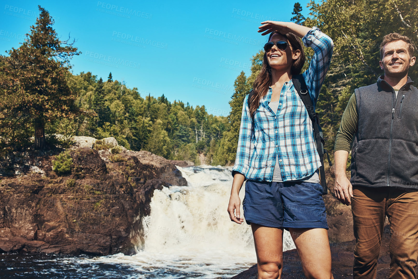 Buy stock photo Shot of a young couple walking next to a rocky river and waterfall