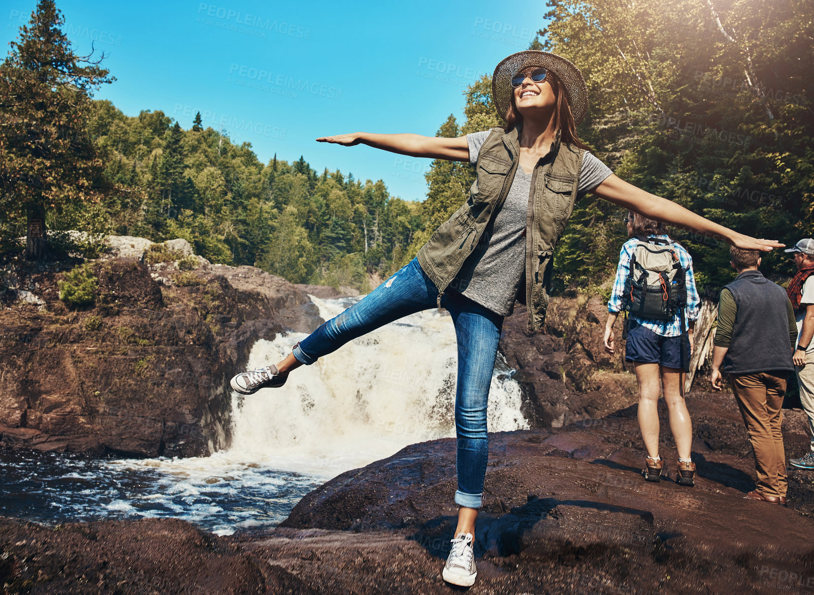 Buy stock photo Shot of a young woman having fun next to a rocky river and waterfall with her friends in the background