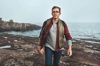 Buy stock photo Shot of a confident middle aged man walking on rocks next to the ocean while wearing a backpack outside during the day