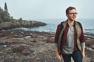 Buy stock photo Shot of a confident middle aged man walking on rocks next to the ocean while wearing a backpack outside during the day