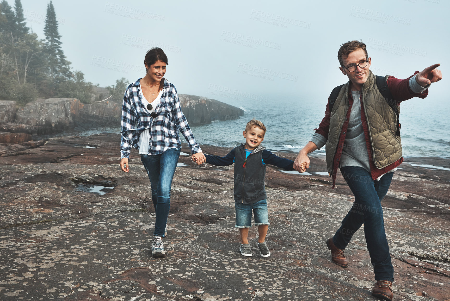 Buy stock photo Shot of a cheerful young family holding hands and walking together next to the ocean outside during the day