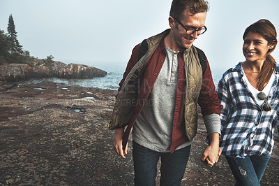 Buy stock photo Shot of a cheerful young couple holding hands and walking together next to the ocean outside during the day