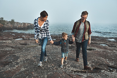 Buy stock photo Shot of a cheerful young family holding hands and walking together next to the ocean outside during the day