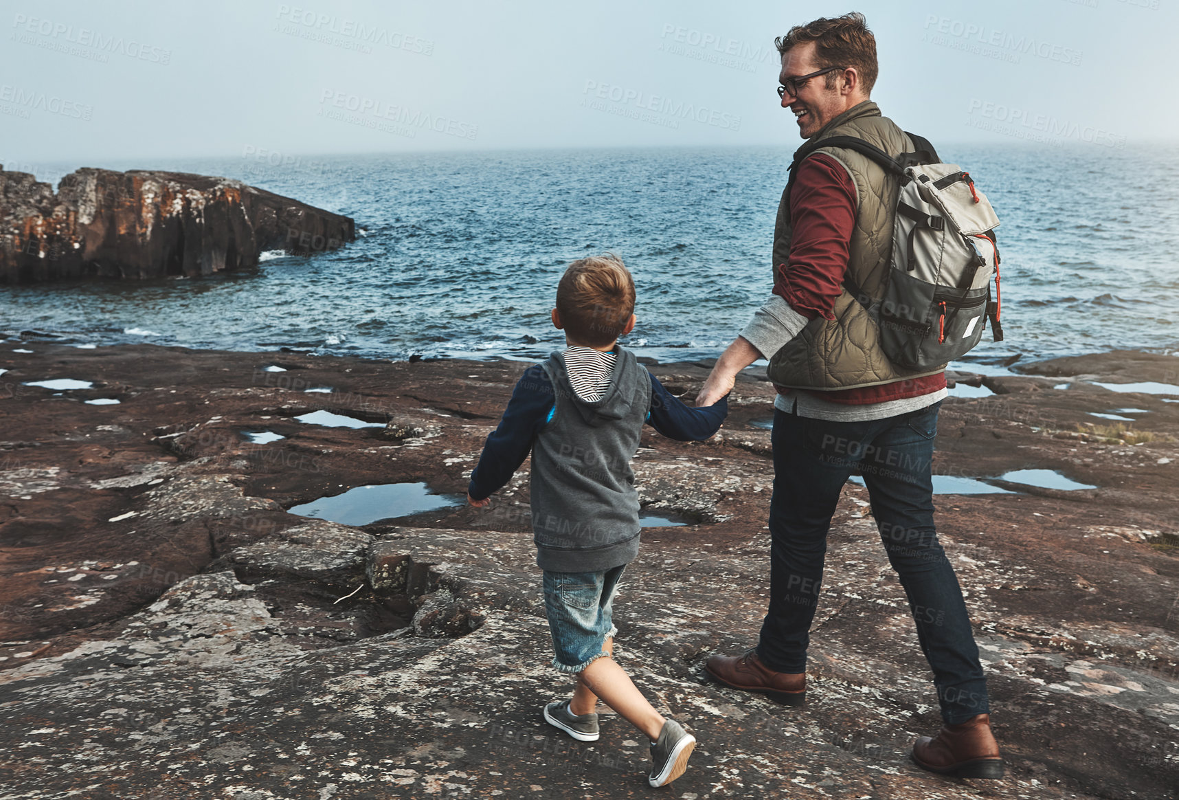 Buy stock photo Rearview shot of a cheerful father and son holding hands while walking next to the ocean outside during the day