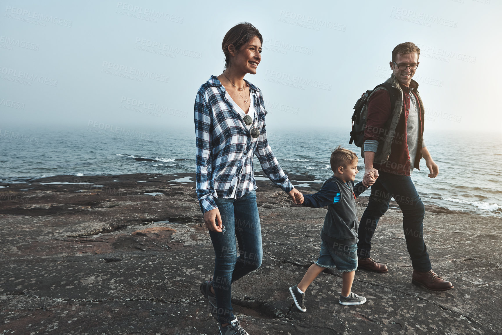 Buy stock photo Shot of a cheerful young family holding hands and walking together next to the ocean outside during the day