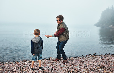 Buy stock photo Rearview shot of a cheerful father and son throwing flat stones into a lake while standing next to the water