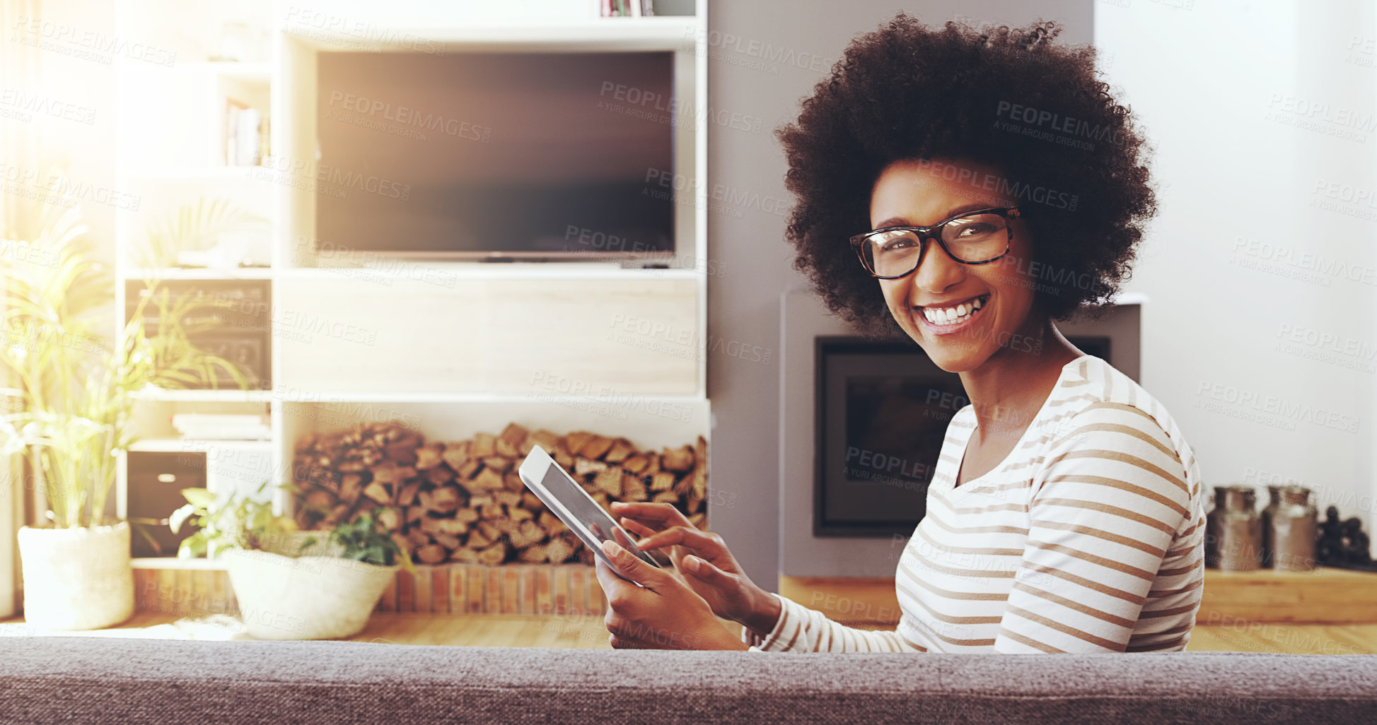 Buy stock photo Portrait of a cheerful young woman wearing glasses while browsing on a digital tablet and seated next to a couch at home
