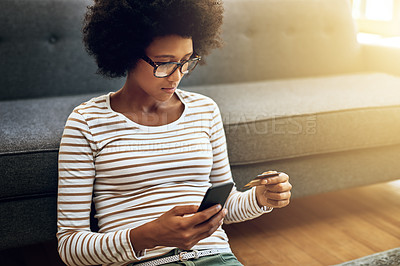 Buy stock photo Shot of a focused young woman doing online banking with her phone while being seated on the floor next to a couch at home
