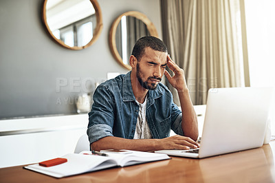 Buy stock photo Shot of a focused young man working on his laptop while holding his head in discomfort at home during the day