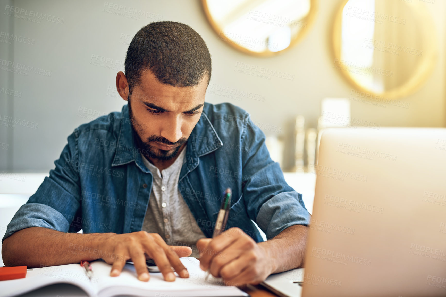 Buy stock photo Shot of a focused young man working on his laptop while making notes in a book behind a table at home during the day