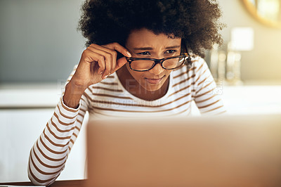 Buy stock photo Shot of a focused young woman working on her laptop while holding her reading glasses at home during the day