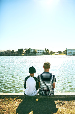 Buy stock photo Rearview shot of two unrecognizable young brothers sitting outside by a lagoon