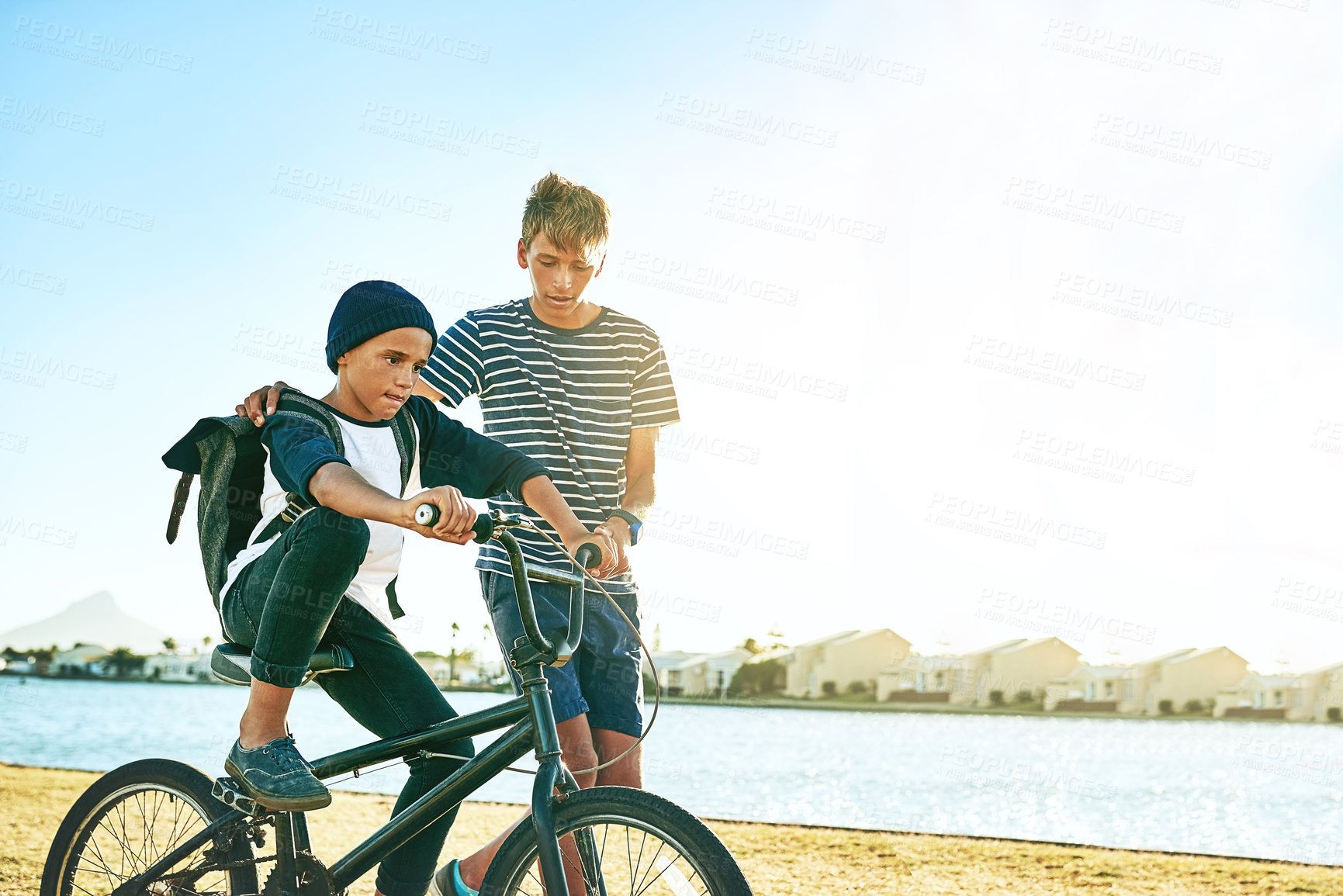 Buy stock photo Cropped shot of a young boy teaching his younger brother how to ride a bike alongside a lagoon