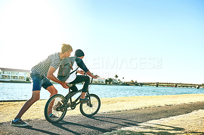 Buy stock photo Full length shot of a young boy teaching his younger brother how to ride a bike alongside a lagoon