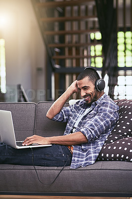 Buy stock photo Shot of a young man using a laptop and headphones on the sofa at home