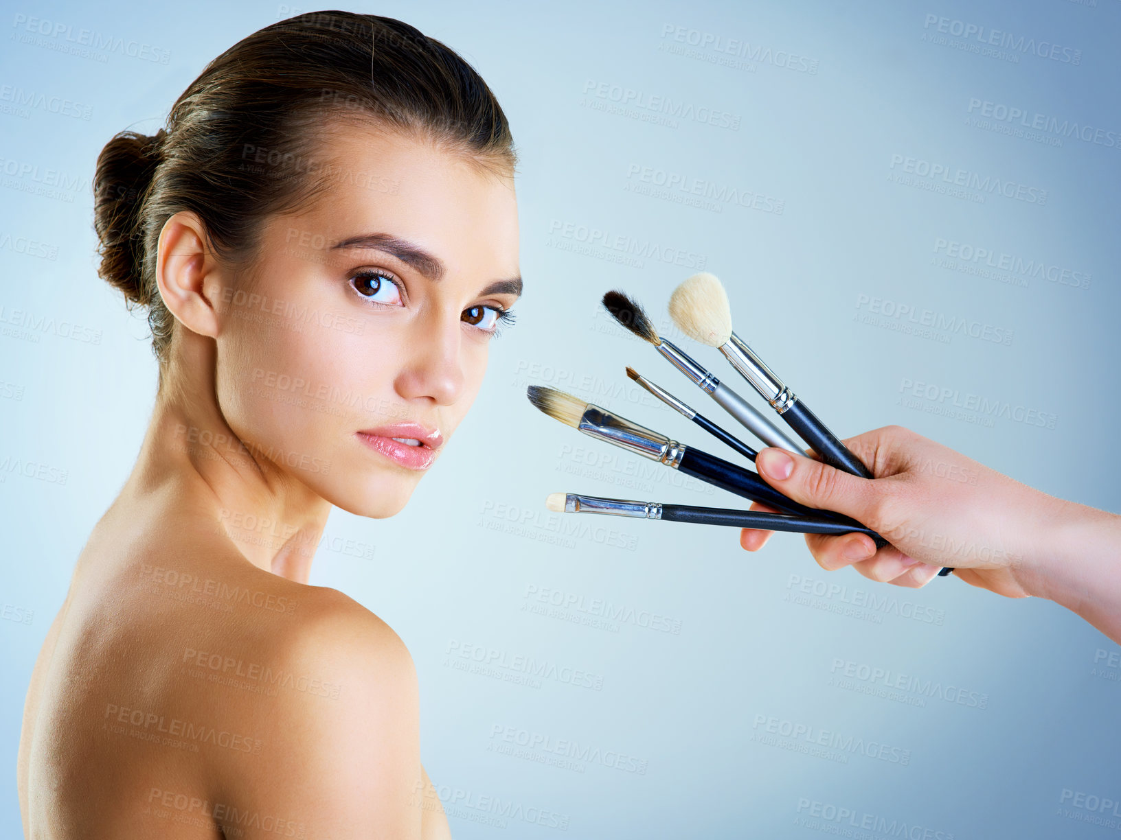 Buy stock photo Studio portrait of a hand holding makeup brushes next to a beautiful young woman against a blue background