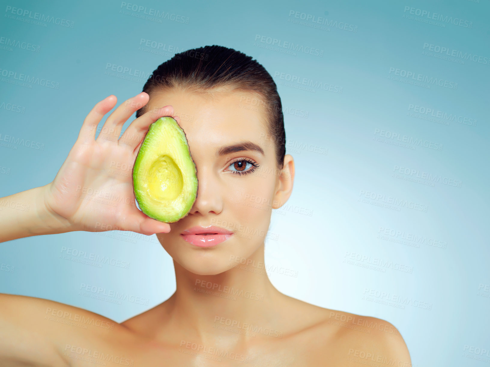 Buy stock photo Studio portrait of a beautiful young woman covering her eye with an avocado against a blue background