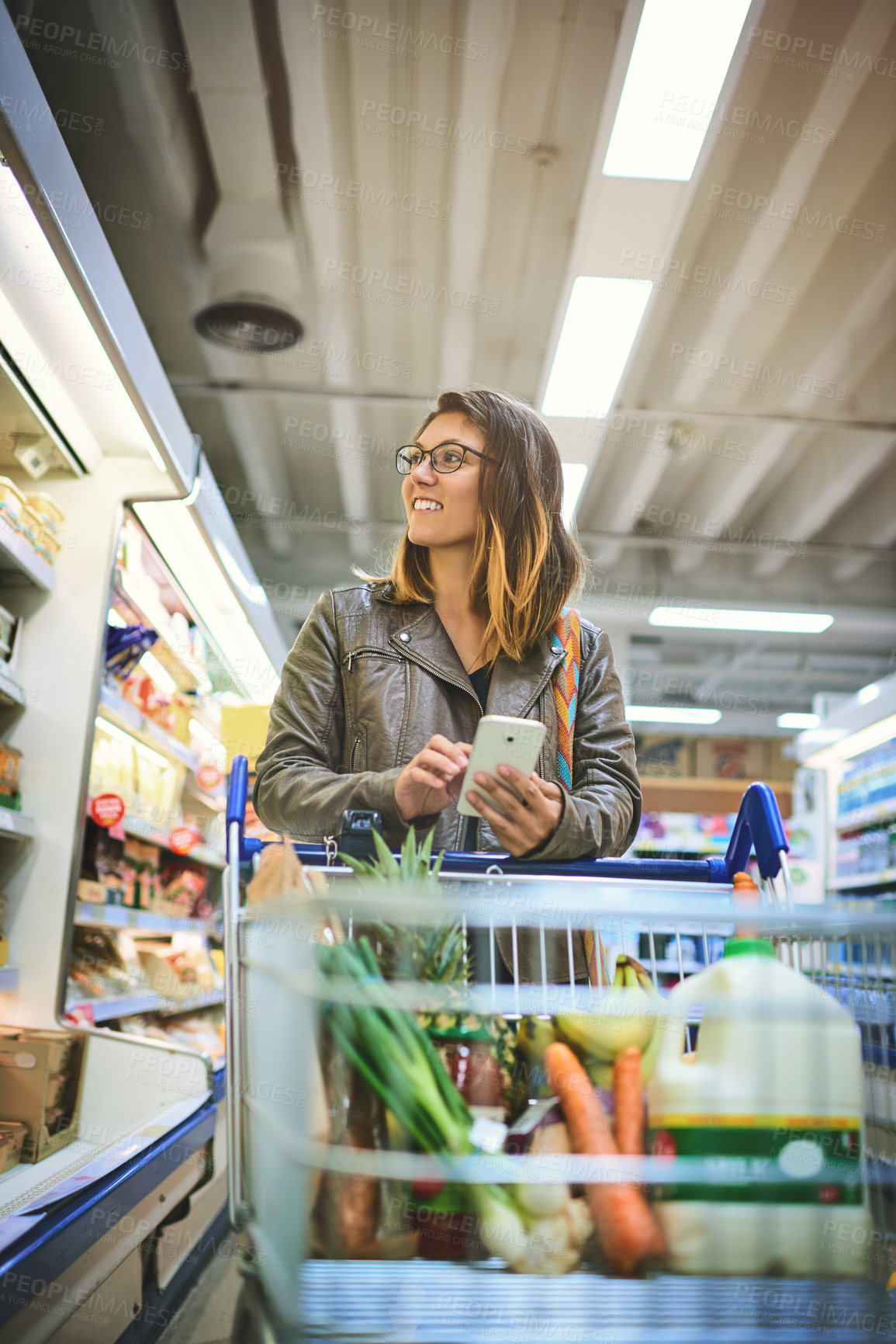 Buy stock photo Shopping, cart and woman with phone in store for healthy food, website and online information. Happy, person and customer with tech in supermarket for grocery list, items and checking ingredients