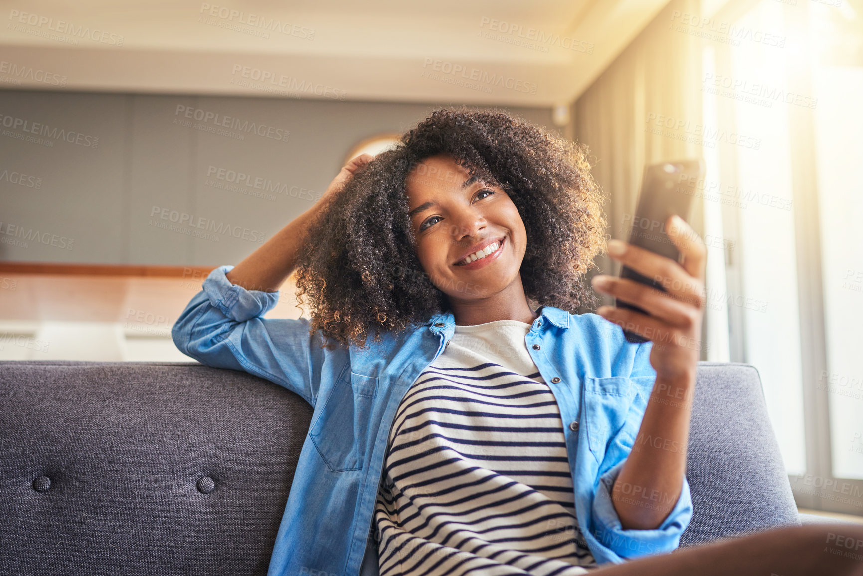 Buy stock photo Shot of a cheerful young woman browsing on her cellphone while being seated on a couch at home during the day