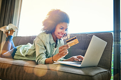 Buy stock photo Shot of a cheerful young woman doing online shopping while lying on a couch at home during the day
