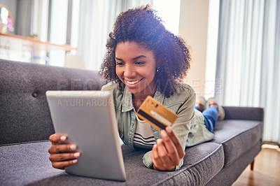 Buy stock photo Shot of a cheerful young woman doing online shopping while lying on a couch at home during the day