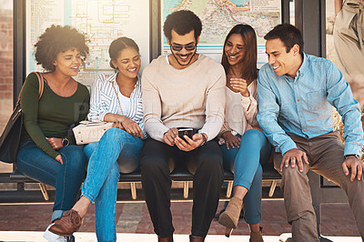 Buy stock photo Shot of a cheerful group of young friends seated together while looking on a mobile phone's screen outside during the day