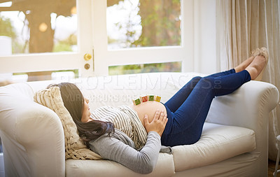 Buy stock photo Shot of an attractive young pregnant woman balancing wooden blocks on her tummy while relaxing on the sofa at home