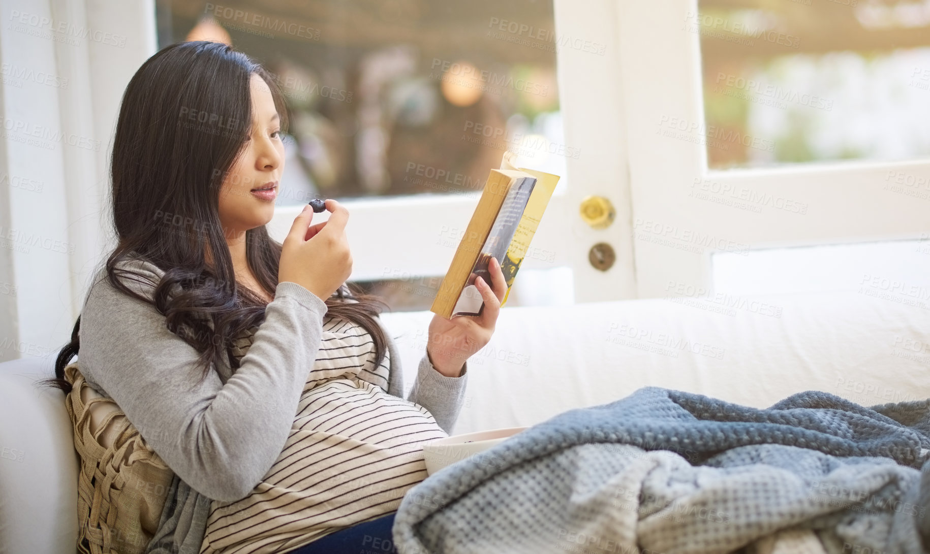 Buy stock photo Cropped shot of an attractive young pregnant woman eating fruit and reading a book while relaxing on the sofa at home