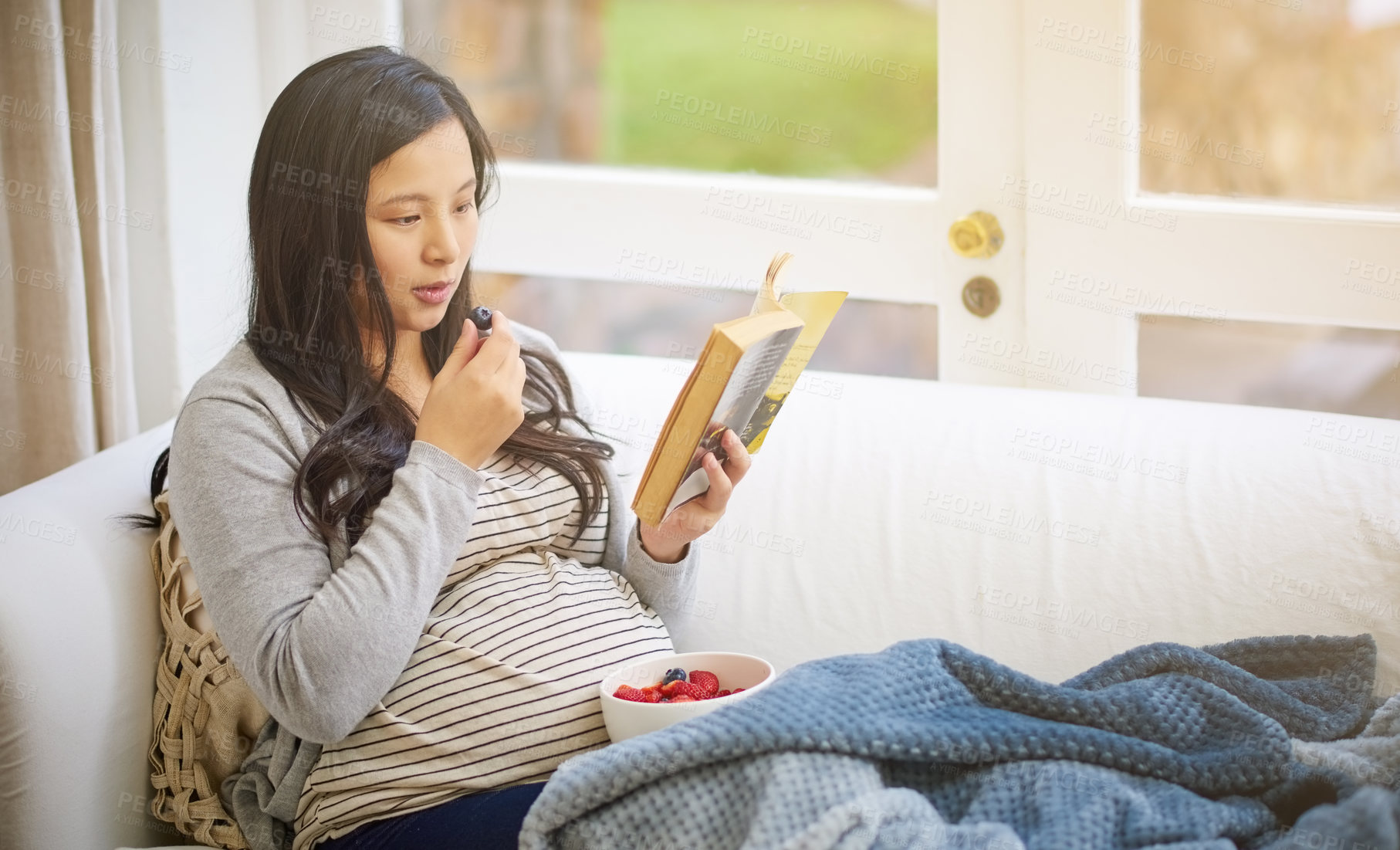 Buy stock photo Cropped shot of an attractive young pregnant woman eating fruit and reading a book while relaxing on the sofa at home