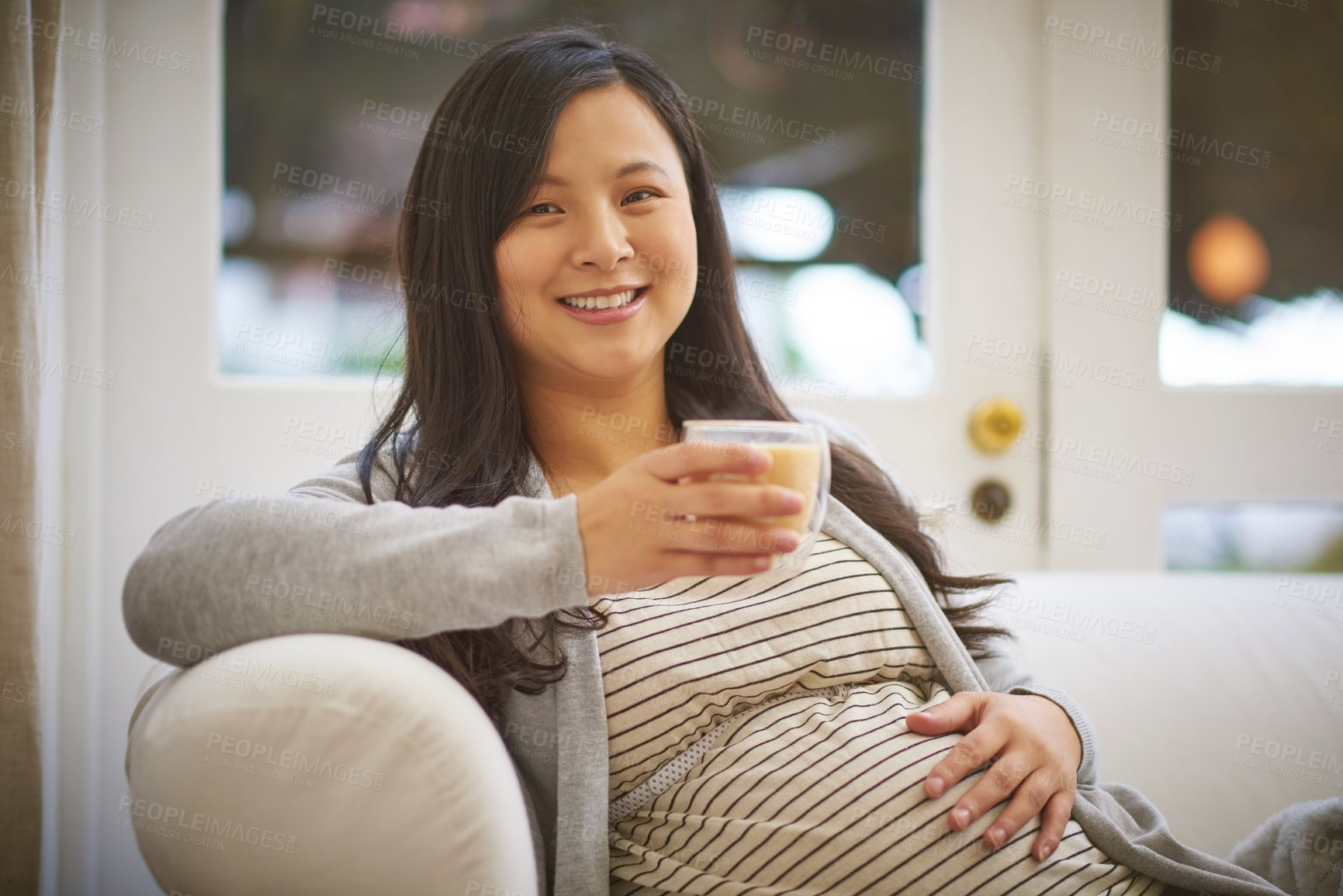 Buy stock photo Portrait of an attractive young pregnant woman drinking an iced coffee while relaxing on the sofa at home