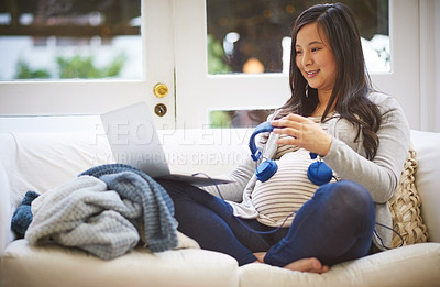 Buy stock photo Full length shot of an attractive young pregnant woman holding headphones to her tummy while relaxing on the sofa at home