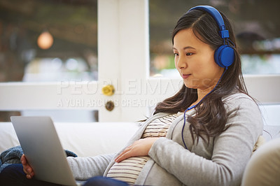 Buy stock photo Shot of a pregnant woman wearing headphones while using a laptop at home