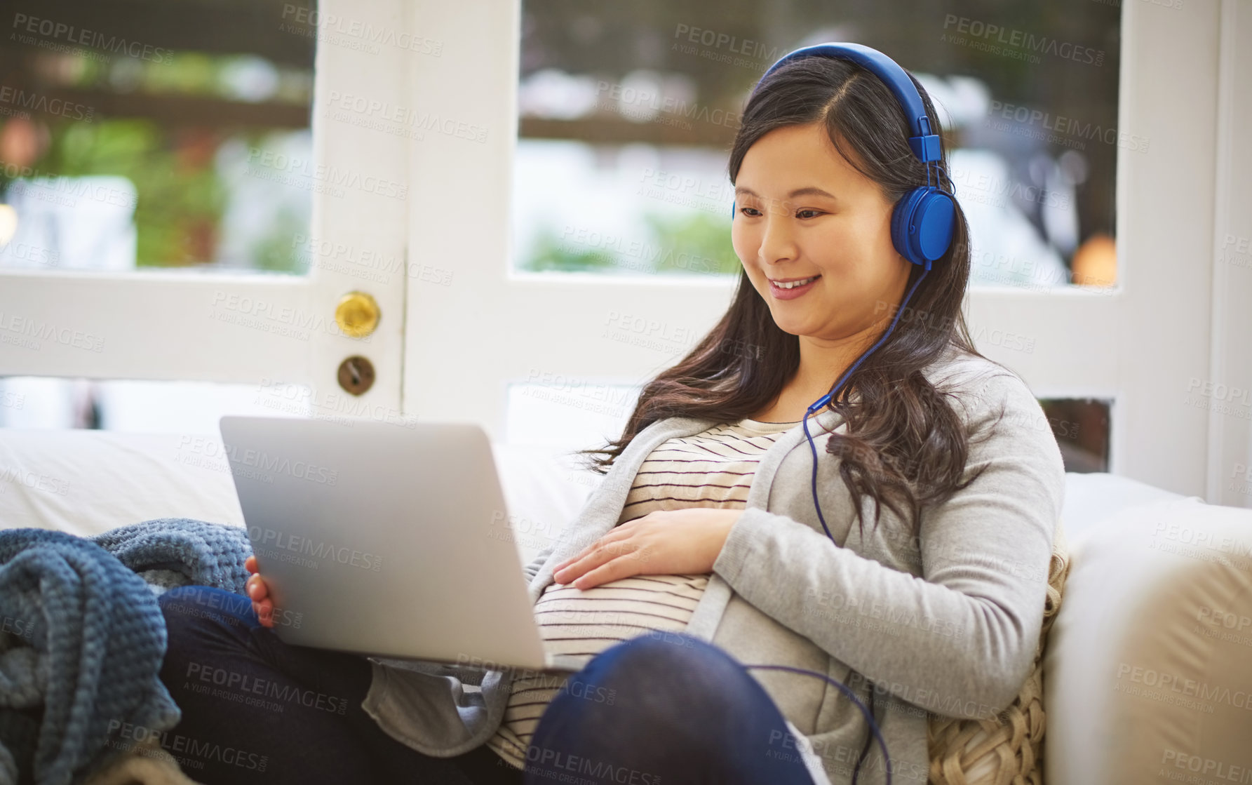 Buy stock photo Shot of a pregnant woman wearing headphones while using a laptop at home