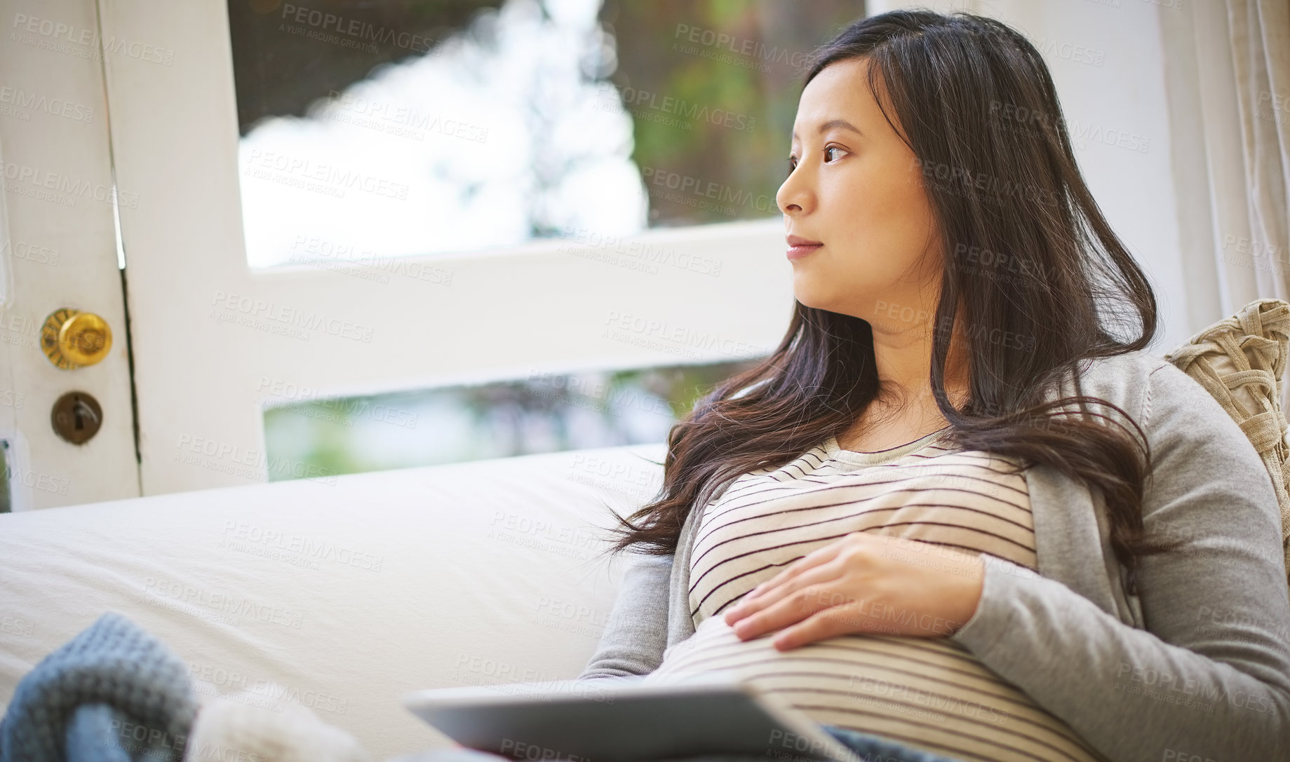 Buy stock photo Shot of a pregnant woman looking thoughtful while using a digital tablet at home