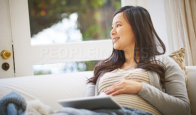 Buy stock photo Shot of a young pregnant woman looking thoughtful while using a digital tablet at home