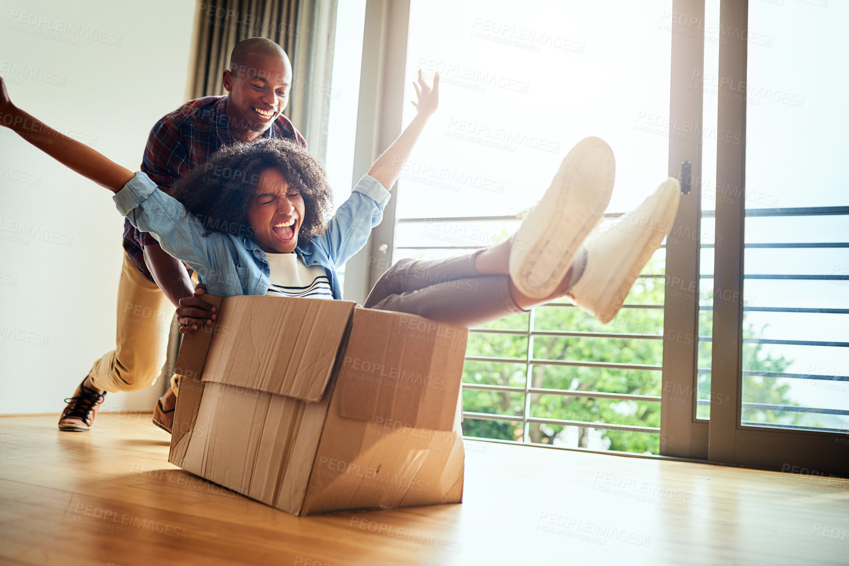Buy stock photo Shot of a cheerful young woman inside of a cardboard box being pushed by her boyfriend at home during the day
