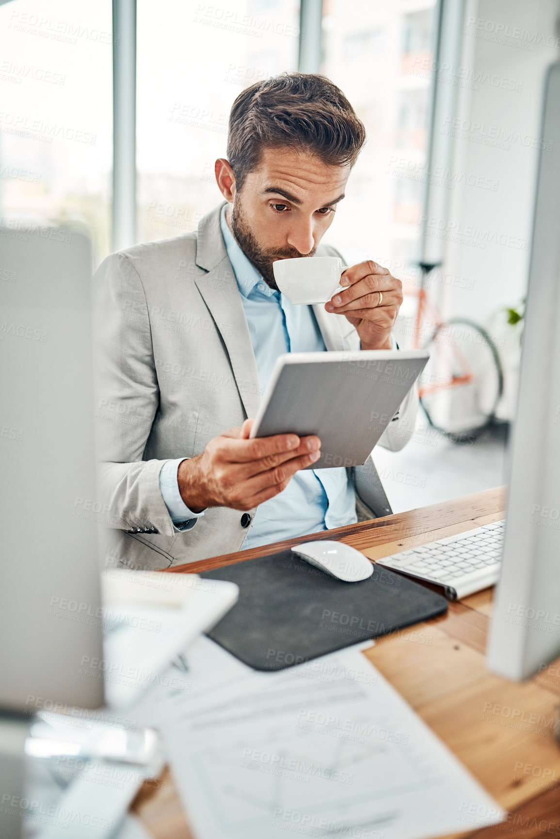Buy stock photo Shot of a handsome young businessman drinking coffee while working on a digital tablet in an office