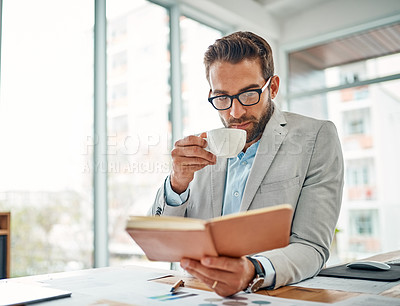 Buy stock photo Shot of a handsome young businessman drinking coffee while reading a book in an office