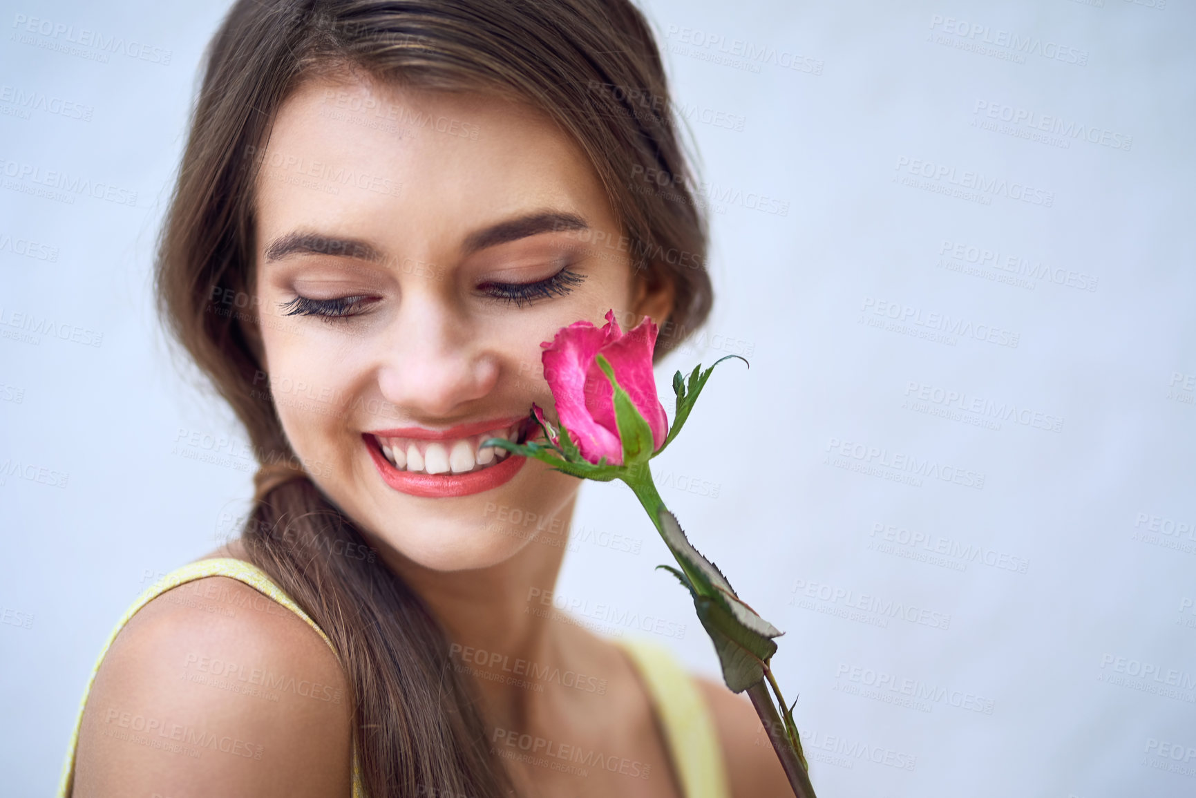 Buy stock photo Studio closeup of a cheerful young woman holding a pink rose next to her face while standing against a grey background