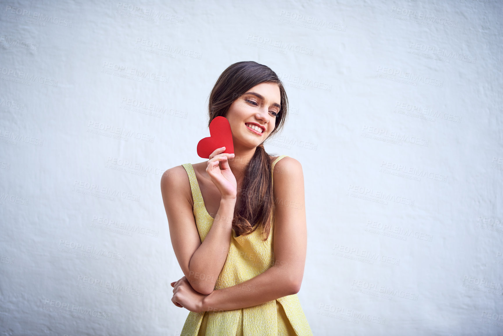 Buy stock photo Studio shot of a cheerful young woman holding a heart shaped piece of paper in her hands while standing against a grey background