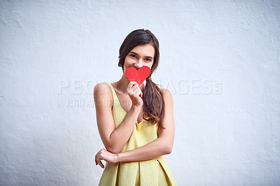 Buy stock photo Studio shot of a cheerful young woman holding a heart shaped piece of paper in her hands while standing against a grey background