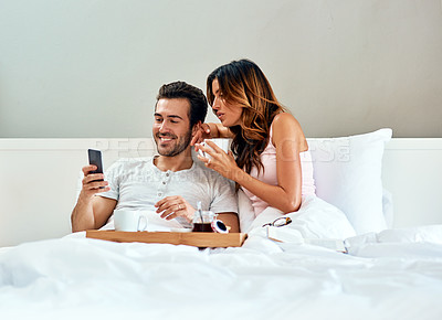 Buy stock photo Shot of a happy young couple using a cellphone while enjoying breakfast in bed together at home